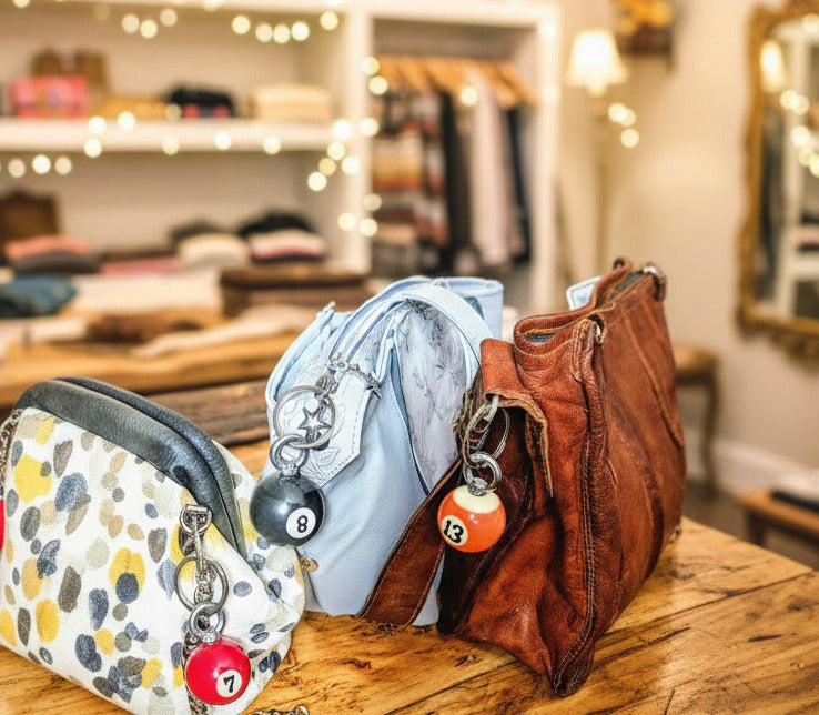 Three handbags displayed on a wooden table inside a boutique: a patterned cream, gray, and yellow pebble bag with red pom-poms, a light blue leather bag with a black 8-ball charm, and a brown leather shoulder bag with a gold-toned 8-ball charm.