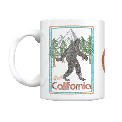 A white ceramic mug featuring a retro-style graphic of a Bigfoot silhouette in front of mountains and pine trees, with the text 'Visit California' in orange and blue.