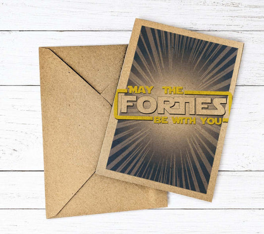 A square kraft paper greeting card with a brown envelope, placed on a white wood plank background. The card features a dark gray and black sunburst design with the text 'MAY THE FORTIES BE WITH YOU' in yellow and beige.