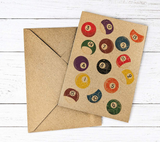 A square kraft paper greeting card with a vintage-style illustration of colorful pool balls, shown with a matching brown envelope on a white wood plank background.