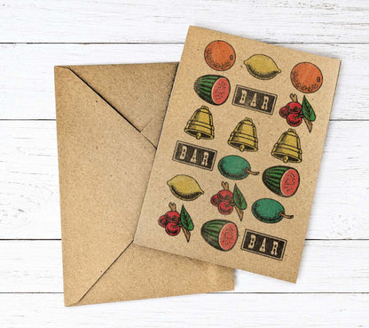 A kraft paper greeting card with vintage-style fruit slot machine logos illustrations of lemons, watermelons, bells, cherries, and the word 'BAR' repeated, shown with a matching kraft envelope on a white wood background.