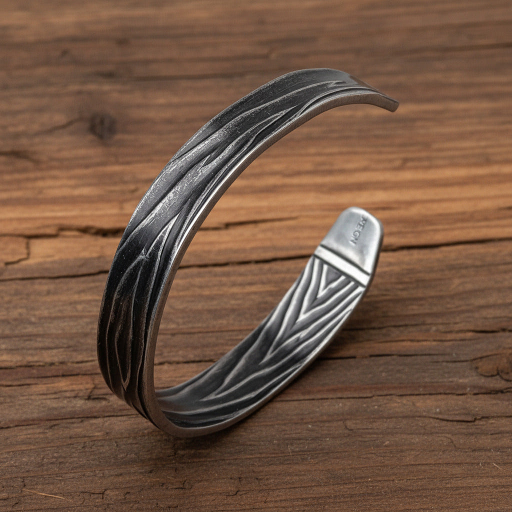 A textured sterling silver cuff bracelet with a wavy, layered pattern and a triangular engraved interior, displayed on a light gray wood background.