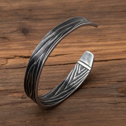 A textured sterling silver cuff bracelet with a wavy, layered pattern and a triangular engraved interior, displayed on a light gray wood background.