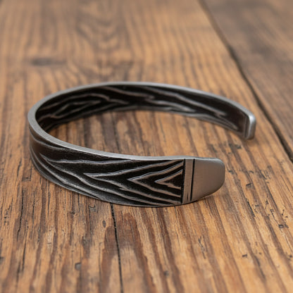 A dark gray metal cuff bracelet with a textured chevron pattern and a polished silver-toned end, displayed on a light, rustic wooden surface.
