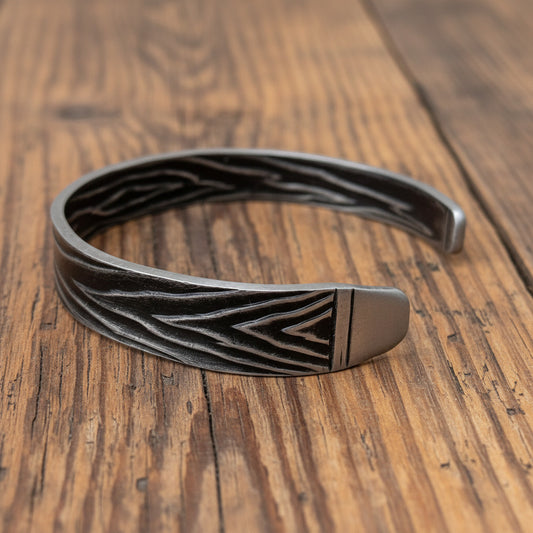 A dark gray metal cuff bracelet with a textured chevron pattern and a polished silver-toned end, displayed on a light, rustic wooden surface.