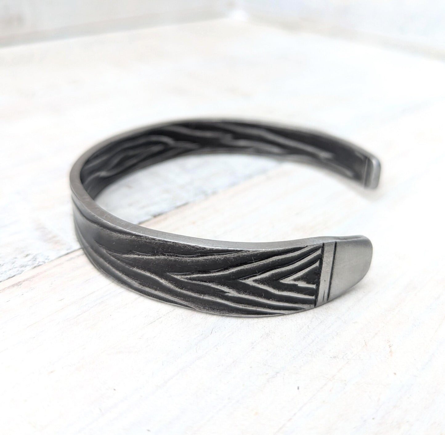 A dark gray metal cuff bracelet with a textured finish and engraved chevron patterns