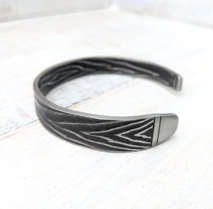A dark gray metal cuff bracelet with a textured finish and engraved chevron patterns