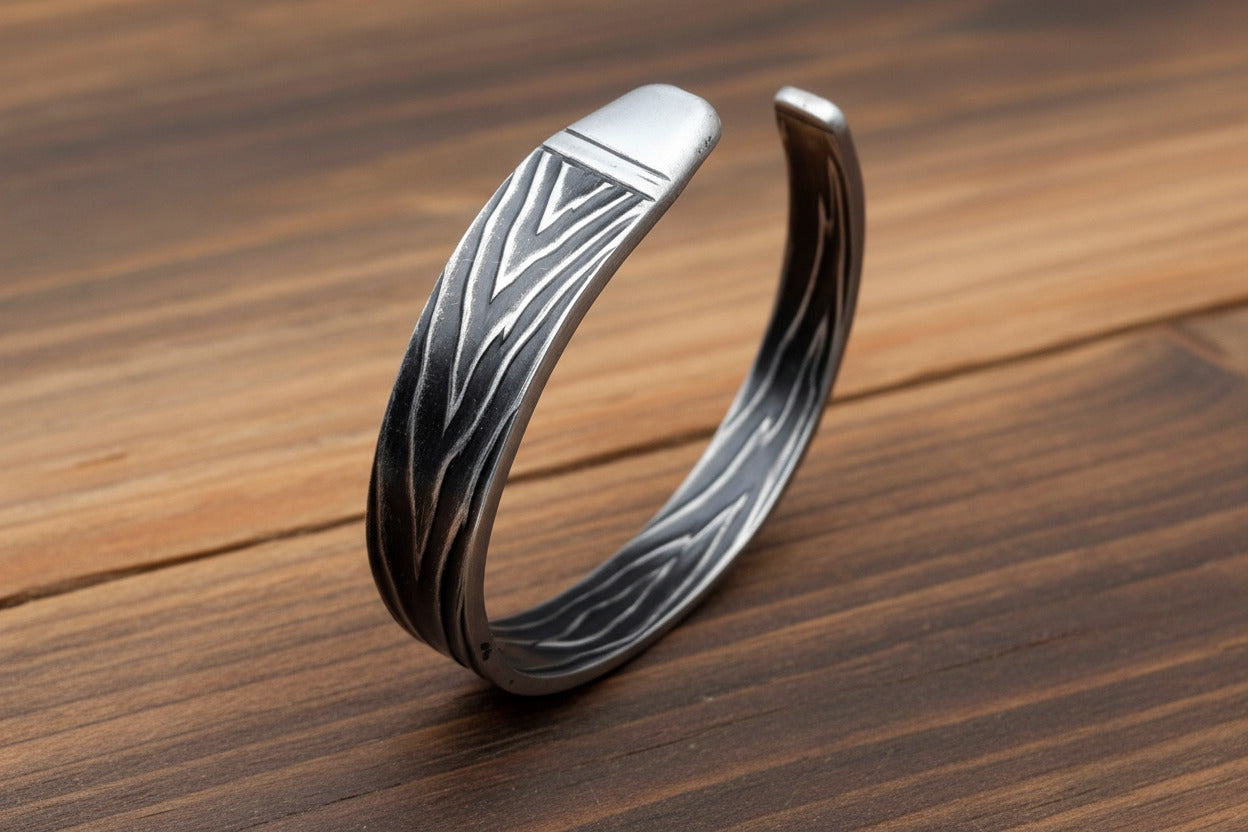 A silver-toned open cuff bracelet with a textured, engraved bark-like pattern, displayed on a light-colored wooden surface.