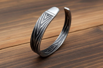 A silver-toned open cuff bracelet with a textured, engraved bark-like pattern, displayed on a light-colored wooden surface.